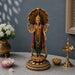 A golden Lord Shiva statue standing on a lotus base with a large open-shell canopy, draped in red and green fabric, displayed on a gray stone table with decorative flowers, a brass lamp, and a bowl of offerings against a dark gray wall.