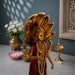 A golden Lord Shiva statue with a multi-headed snake canopy, standing on a gray stone surface next to a brass diya, a pink lotus bowl, and a vase of white flowers against a dark blue-gray wall.