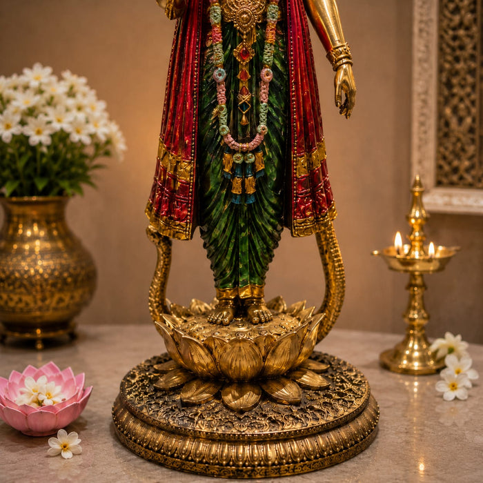 A decorative golden statue of a Hindu deity standing on a lotus pedestal, draped in red and green garments, displayed indoors on a stone surface with flowers and a brass oil lamp in the background.