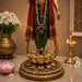 A decorative golden statue of a Hindu deity standing on a lotus pedestal, draped in red and green garments, displayed indoors on a stone surface with flowers and a brass oil lamp in the background.