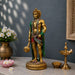 A golden Lord Hanuman statue adorned with green and red draped fabric, standing on a decorative base and holding a mace, displayed on a marble tabletop with a brass lamp, flowers, and a dark gray wall background.