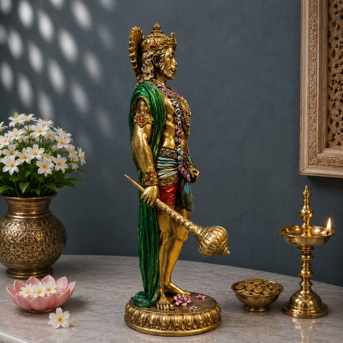 A golden Lord Hanuman statue standing on a decorated base, holding a mace, styled on a marble tabletop with a gold oil lamp, lotus flowers, and a vase of white flowers against a dark gray wall.