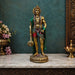 A golden Lord Hanuman statue with green and red draped fabric, standing on a marble mantelpiece against a dark teal textured wall.