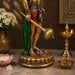 A decorative golden statue of a Hindu deity standing on a lotus base, styled on a tabletop with flowers, a lit brass diya, and a small bowl in a warm indoor setting.