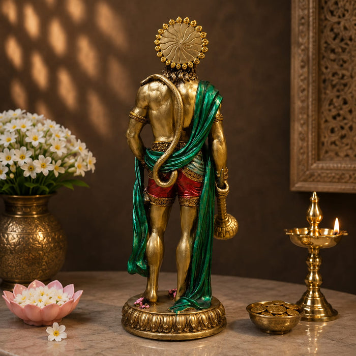 A golden Lord Hanuman statue shown from the back, draped in a green shawl and wearing red shorts, standing on a marble surface with decorative flowers, a brass lamp, and a carved wall in the background.