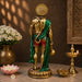 A golden Lord Hanuman statue shown from the back, draped in a green shawl and wearing red shorts, standing on a marble surface with decorative flowers, a brass lamp, and a carved wall in the background.