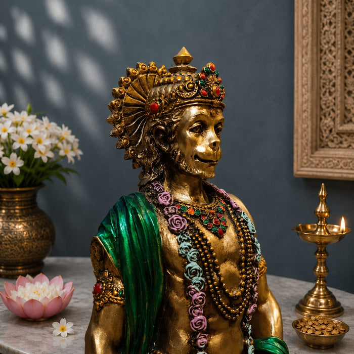 A golden Hanuman statue adorned with colorful floral garlands and a green shawl, displayed on a marble surface with a vase of white flowers, a pink lotus, and a brass oil lamp against a dark blue-gray wall.