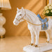 A white ceramic horse figurine with a light blue saddle and gold trim, standing on a decorative base in a warmly lit indoor setting.