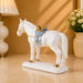 A white ceramic horse figurine with gold and light blue saddle details, displayed on a beige tabletop with a warm-toned lamp and flowers in the background.