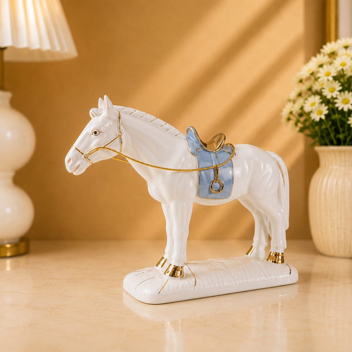 A white ceramic horse figurine standing on a matching base, decorated with a light blue saddle and gold accents, displayed on a beige tabletop with a lamp and flowers in the background.