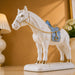 A white ceramic horse figurine with a light blue saddle and gold trim, standing on a matching white base against a warm beige background.