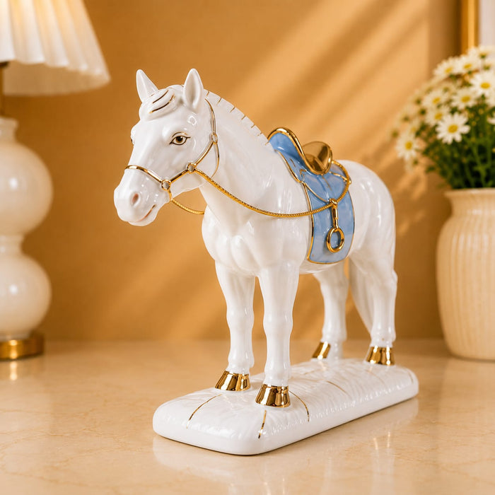 A white ceramic horse figurine with gold trim and a light blue saddle, displayed on a beige tabletop with a warm-toned lamp and flowers in the background.