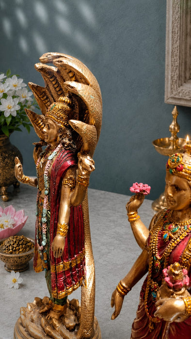 A close-up of a decorative golden Hindu deity statue, likely Shiva Nataraja, with a multi-headed snake canopy and red draped garments, shown alongside another similar statue in an indoor setting.