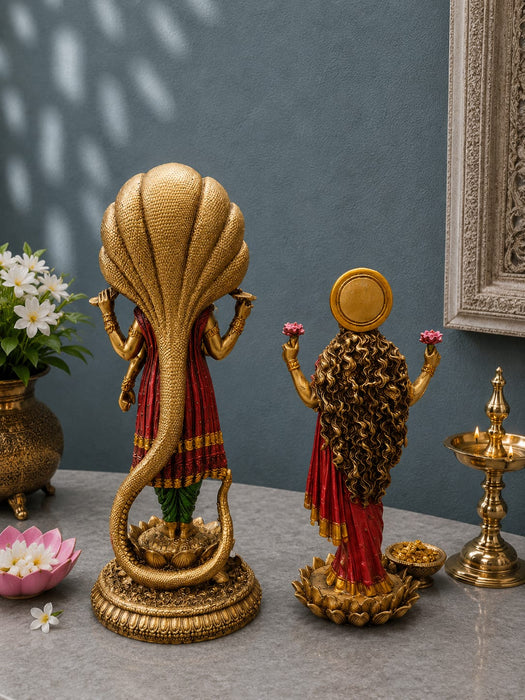 Two ornate gold-toned decorative statues displayed on a tabletop against a muted blue-gray wall. The larger left statue depicts a serpent-like figure draped in red and green, while the smaller right statue shows a female figure with long dark hair, red drapery, and pink flower accents.