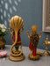 Two ornate gold-toned decorative statues displayed on a tabletop against a muted blue-gray wall. The larger left statue depicts a serpent-like figure draped in red and green, while the smaller right statue shows a female figure with long dark hair, red drapery, and pink flower accents.
