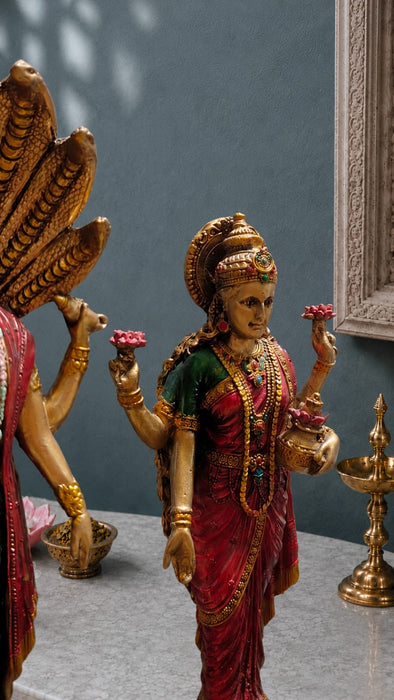 A detailed golden statue of a Hindu goddess, likely Lakshmi, adorned in a red and green sari with gold jewelry, holding pink lotus flowers, displayed next to a bronze Nandi bull statue on a gray surface.