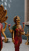 A detailed golden statue of a Hindu goddess, likely Lakshmi, adorned in a red and green sari with gold jewelry, holding pink lotus flowers, displayed next to a bronze Nandi bull statue on a gray surface.