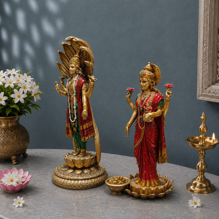 Two ornate gold-toned Hindu deity statues displayed on a gray tabletop against a slate-blue wall, surrounded by decorative props including a brass lamp, flowers, and a small bowl.