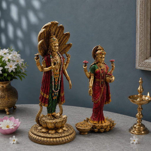 Two ornate gold-toned Hindu deity statues displayed on a gray tabletop against a muted blue-gray wall. The larger statue on the left depicts a multi-headed serpent-decorated figure on a lotus base, while the smaller statue on the right shows a female deity in red and green drapery holding pink flowers.