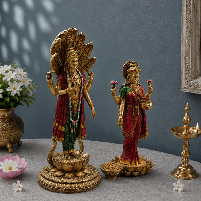 Two ornate gold-toned Hindu deity statues displayed on a gray tabletop against a muted blue-gray wall. The larger statue on the left depicts a multi-headed serpent-decorated figure on a lotus base, while the smaller statue on the right shows a female deity in red and green drapery holding pink flowers.