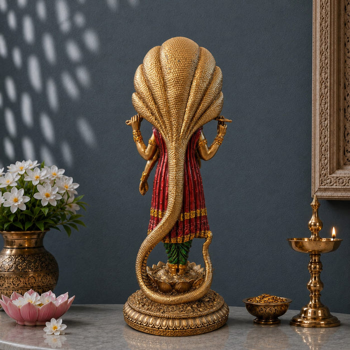 A decorative brass Lord Shani statue with a golden serpent body and a red draped shroud, displayed on a marble surface with a dark gray wall, flowers, and brass oil lamps in the background.