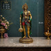 Gold-toned Hindu deity statue holding a mace, displayed on a marble surface with overlaid size measurements (26 cm by 26 cm by 9 cm) against a dark teal wall with decorative flowers and a brass lamp.